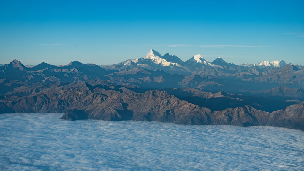 從高空俯瞰不丹的喜馬拉雅山脈,雪白的山峰聳立於藍天之下,與層層疊疊的山脈形成壯觀景象。下方雲海翻騰,營造出如仙境般的視覺效果,顯示不丹作為高山王國的地理奇觀。