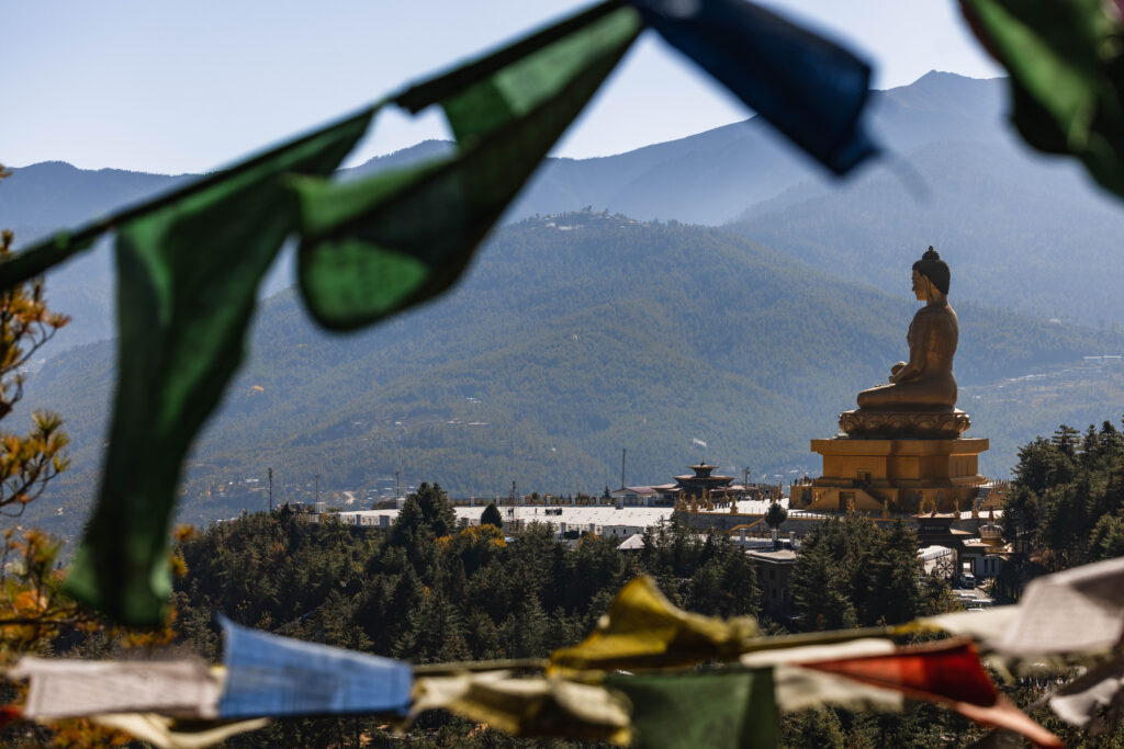 這張圖片捕捉了不丹著名的「露天釋迦牟尼佛像」（Buddha Dordenma），前景為迎風飄揚的五色經幡，遠方則是森林與山巒交錯的壯闊景色。這座金色佛像俯瞰首都廷布（Thimphu），象徵著和平與庇佑。