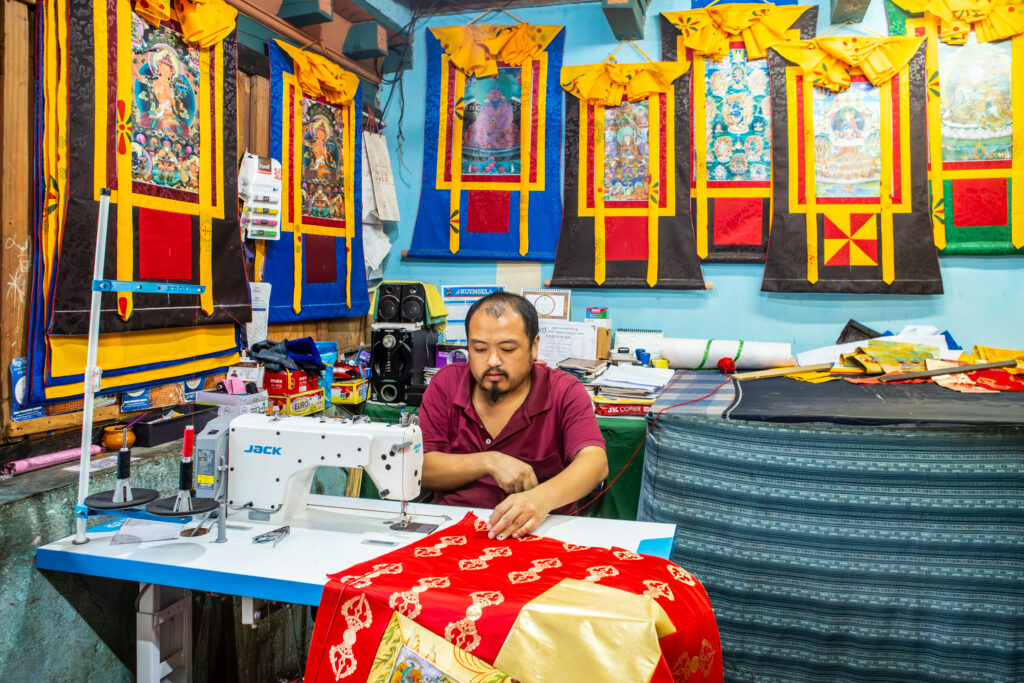 一位工匠坐在縫紉機前，專注地縫製一幅紅色的唐卡（Thangka）。背景牆上掛滿了不同的佛教唐卡，色彩鮮豔，金色裝飾細緻，展現不丹傳統工藝的精湛技術。