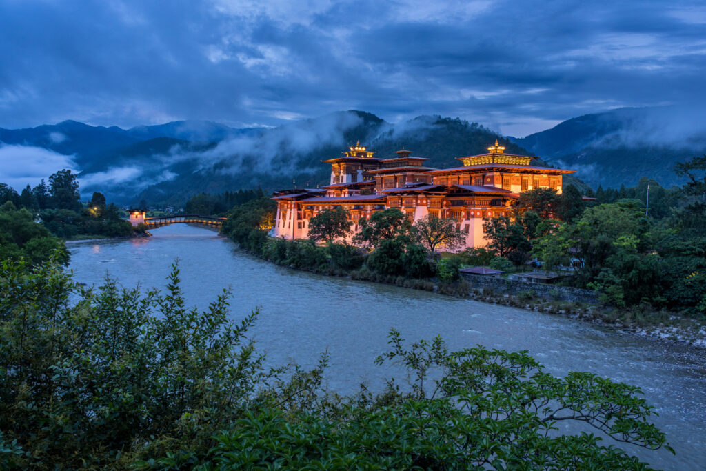 夜色中，普那卡宗（Punakha Dzong）華麗的燈光倒映在河面上，周圍環繞著層層青山與霧氣，展現出不丹最壯觀的宗堡之一。