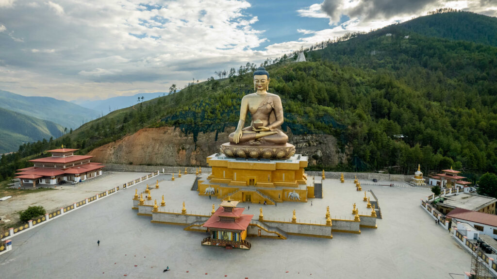 這張圖片展示了不丹廷布（Thimphu）的釋迦牟尼佛像（Buddha Dordenma），這座巨大的青銅佛像鍍金而成，高達51.5公尺，是不丹的地標之一。佛像周圍的廣場擺放著金色的佛塔與雕像，背景則是鬱鬱蔥蔥的山林，景色壯麗且寧靜。