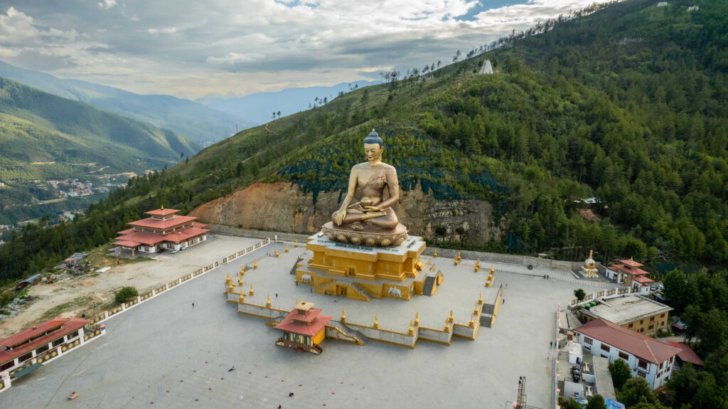 從空中俯瞰廷布的釋迦牟尼佛像，整座佛像矗立在山丘之上，環繞著修行殿堂與多座金色雕像，遠方可見廷布市區與層疊的群山，呈現出不丹獨特的宗教與自然交融的景觀。