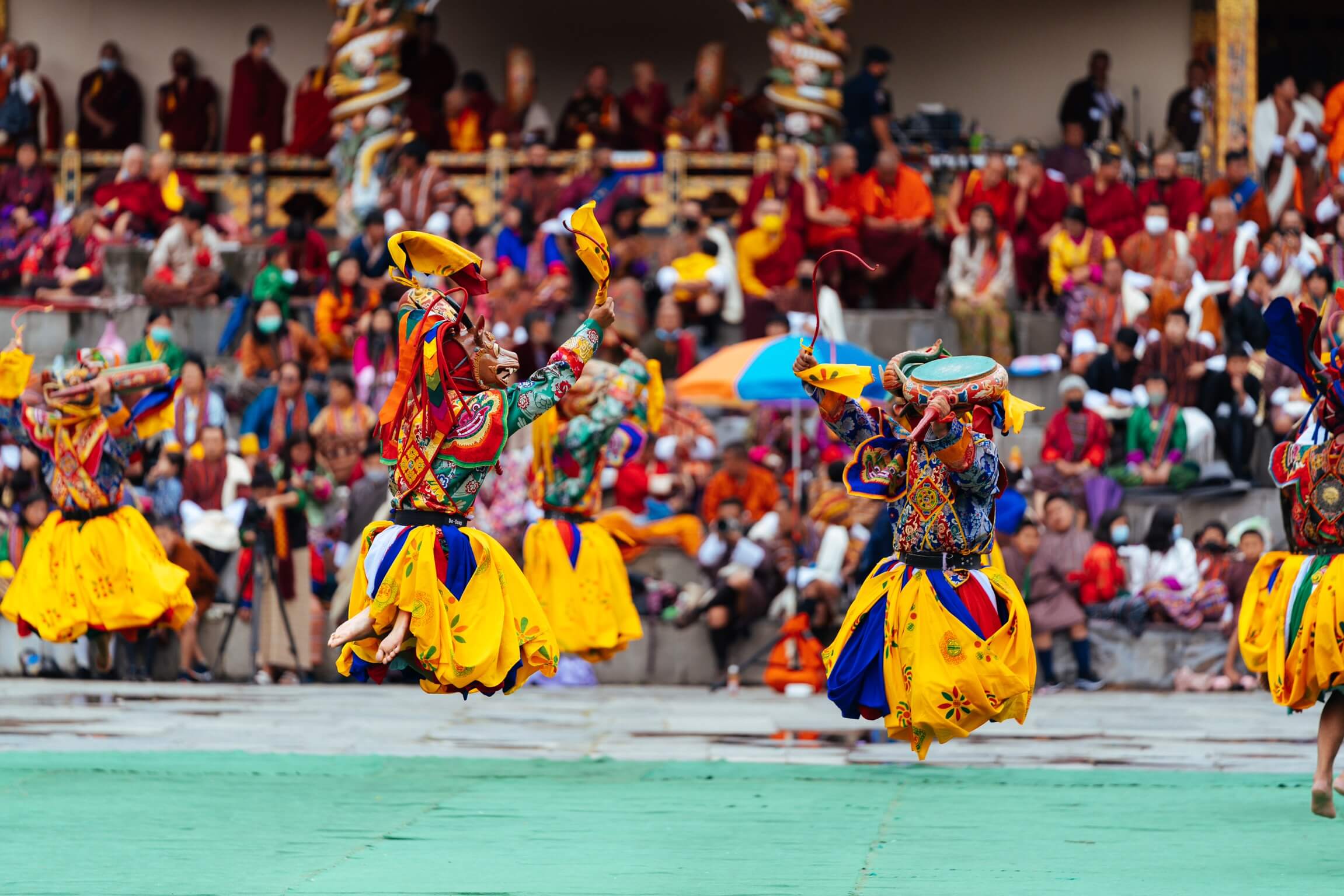 圖片拍攝於不丹宗教節慶期間，舞者身穿鮮豔華麗的傳統服飾，頭戴精緻面具，正躍起舞動，展現不丹宗教與文化的深厚底蘊。背景中可見僧侶與民眾圍觀，場面熱鬧非凡，充分展現不丹節慶的獨特魅力與活力。