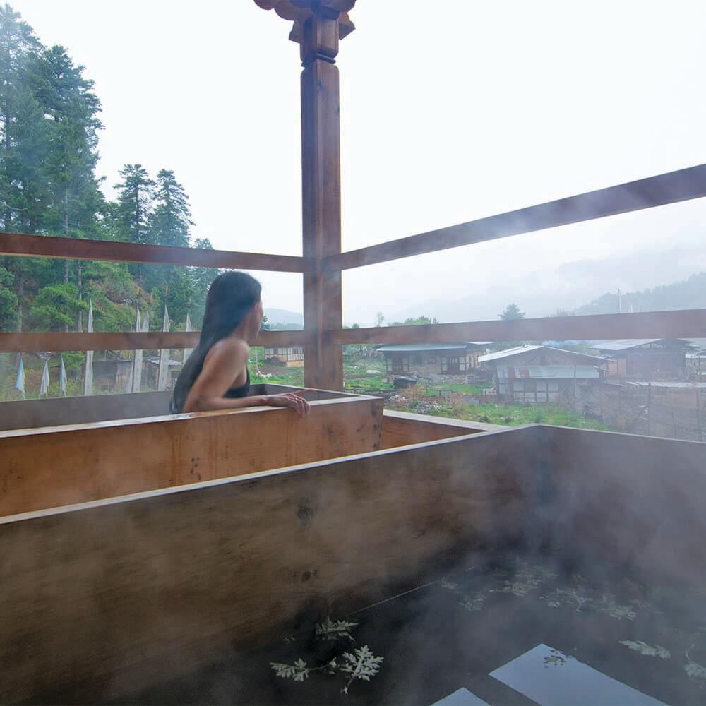 不丹熱石浴（Hot Stone Bath）是一種獨特的傳統療法，使用加熱至高溫的河石投入木製浴槽中，使水溫升高，並融入當地藥草，據說有助於放鬆身心並促進血液循環。圖中可見一位旅人沉浸在熱氣騰騰的木製浴池中，遠望著寧靜的鄉村景色，享受不丹大自然帶來的療癒體驗。