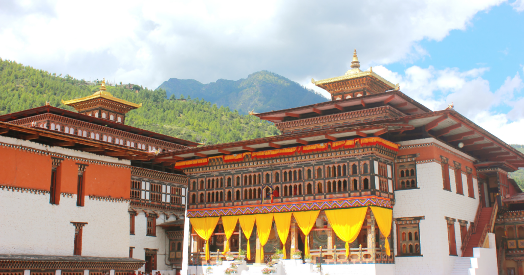 這張照片展示了不丹首都廷布（Thimphu）的札西確宗，這座壯觀的傳統建築擁有白色與紅色相間的牆面，屋頂則裝飾著金色塔尖，象徵不丹的宗教與政治中心。前方懸掛著黃色布幔，表示當地重要的宗教或官方儀式即將舉行，背景是青翠的山巒，突顯札西確宗與自然環境的和諧融合。