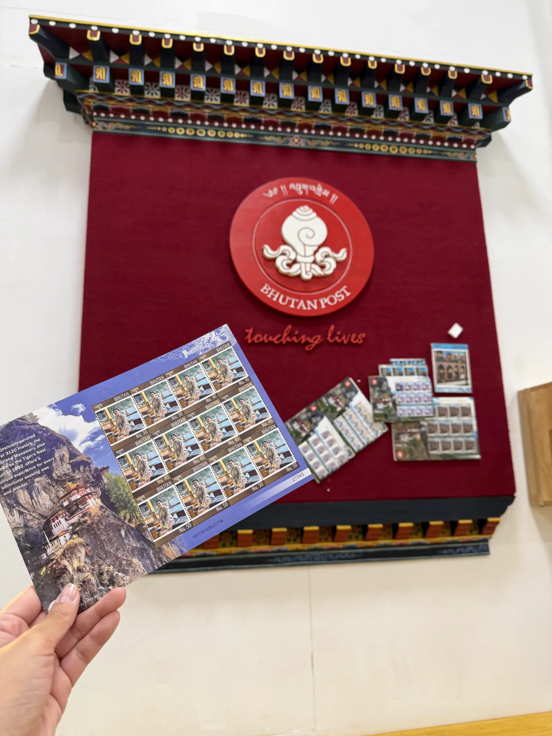 該圖片為不丹中央郵局(Bhutan Post)的參觀紀錄。手持的主視覺是一整版精美的限量不丹郵票,左側印有建在懸崖峭壁上的虎穴寺壯麗風光。背景為郵局內部的深紅色展示牆,牆頂帶有精緻的不丹傳統木雕彩繪裝飾,中央懸掛著圓形的「BHUTAN POST」官方標誌,右側則展示著各式珍藏的歷史郵票套組,呈現出不丹作為「郵票王國」的獨特文化美學。