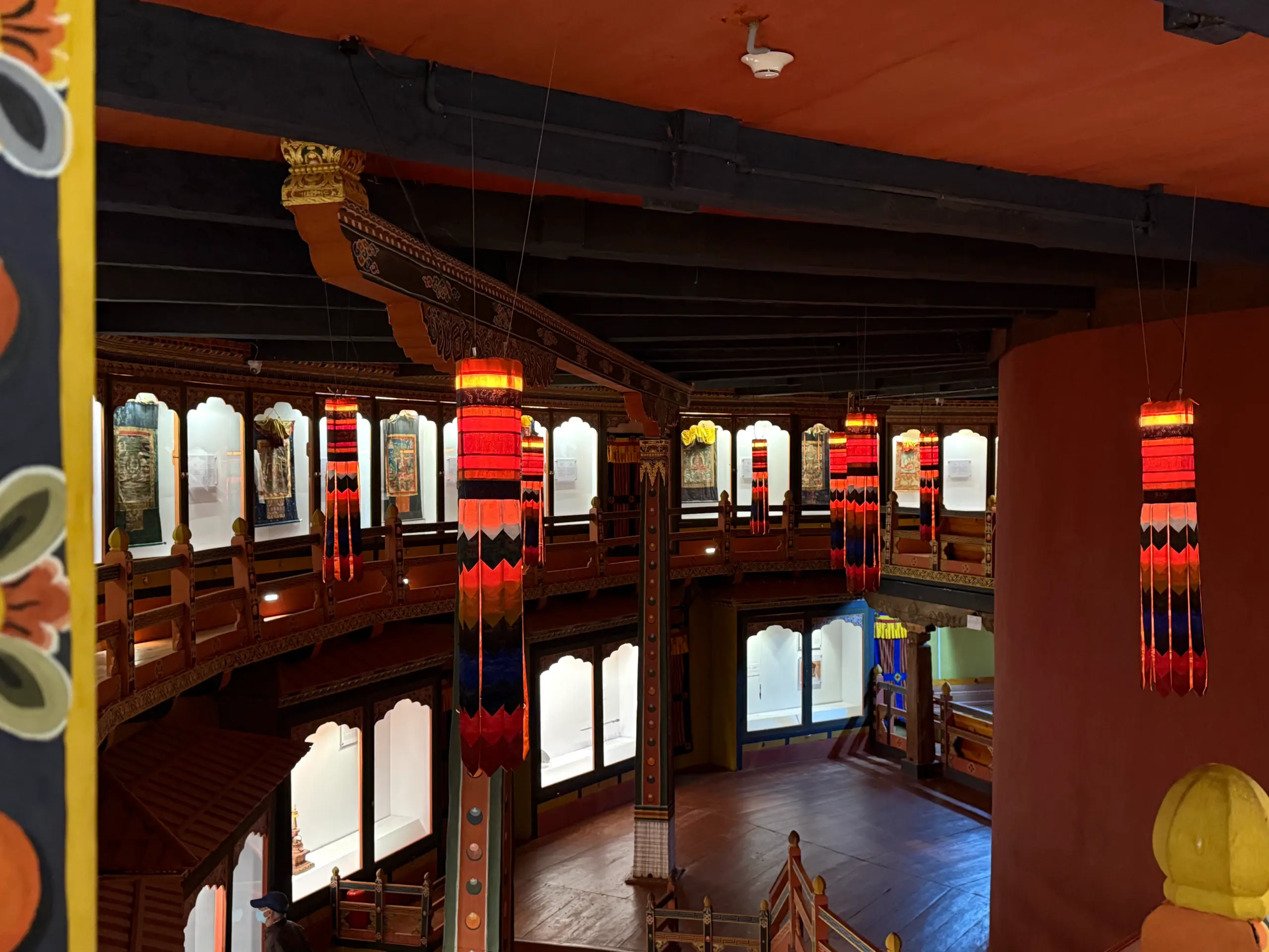 該圖片展示了位於帕羅的不丹國家博物館(National Museum of Bhutan)內部展廳。展間呈現獨特的圓弧形迴廊結構,厚實的土紅色牆面搭配黑色的木質天花板與橫樑。樑柱上懸掛著多盞色彩鮮豔、極具藏傳佛教特色的長條狀織錦法幡(筒狀幡);兩側則整齊排列著點亮燈光的透明玻璃展櫃,內部陳列著唐卡、佛像等不丹歷史上的珍貴宗教文物,散發出神祕且肅穆的歷史氣息。