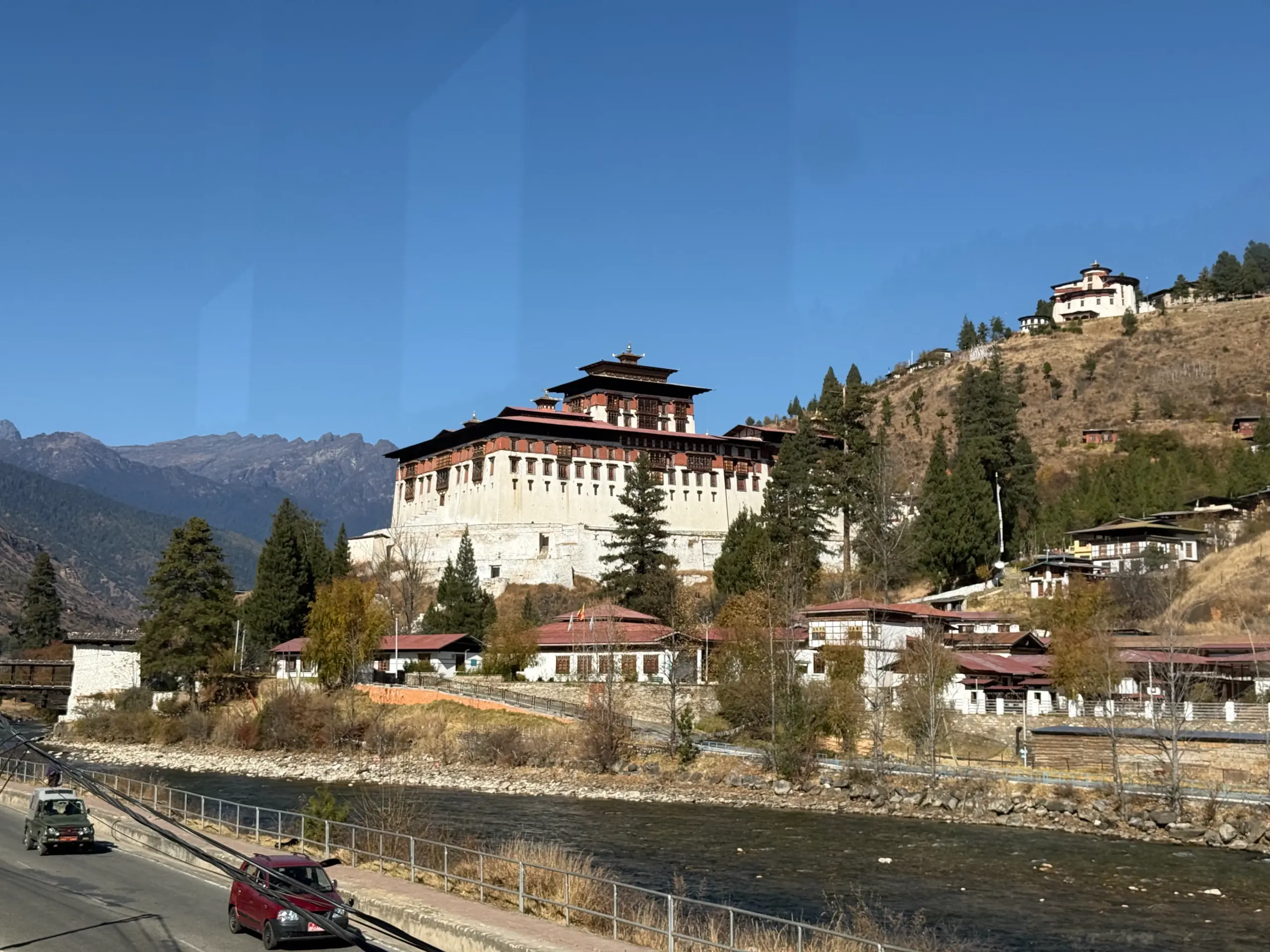 該圖片展示了高聳於山坡上的「帕羅宗(Paro Dzong,又稱仁朋堡)」。主體建築是一座巨大的白色藏式城堡,牆面開有整齊細緻的傳統木窗,頂端設有層層紅瓦屋頂。下方清澈的河流依山流過,河岸兩側分佈著帶有不丹傳統紅瓦屋頂的白色民居小鎮。從行進的車窗向外望去,雄偉的宗堡在藍天與枯黃山色間拔地而起,展現出堅固不摧的宗教要塞氣勢。